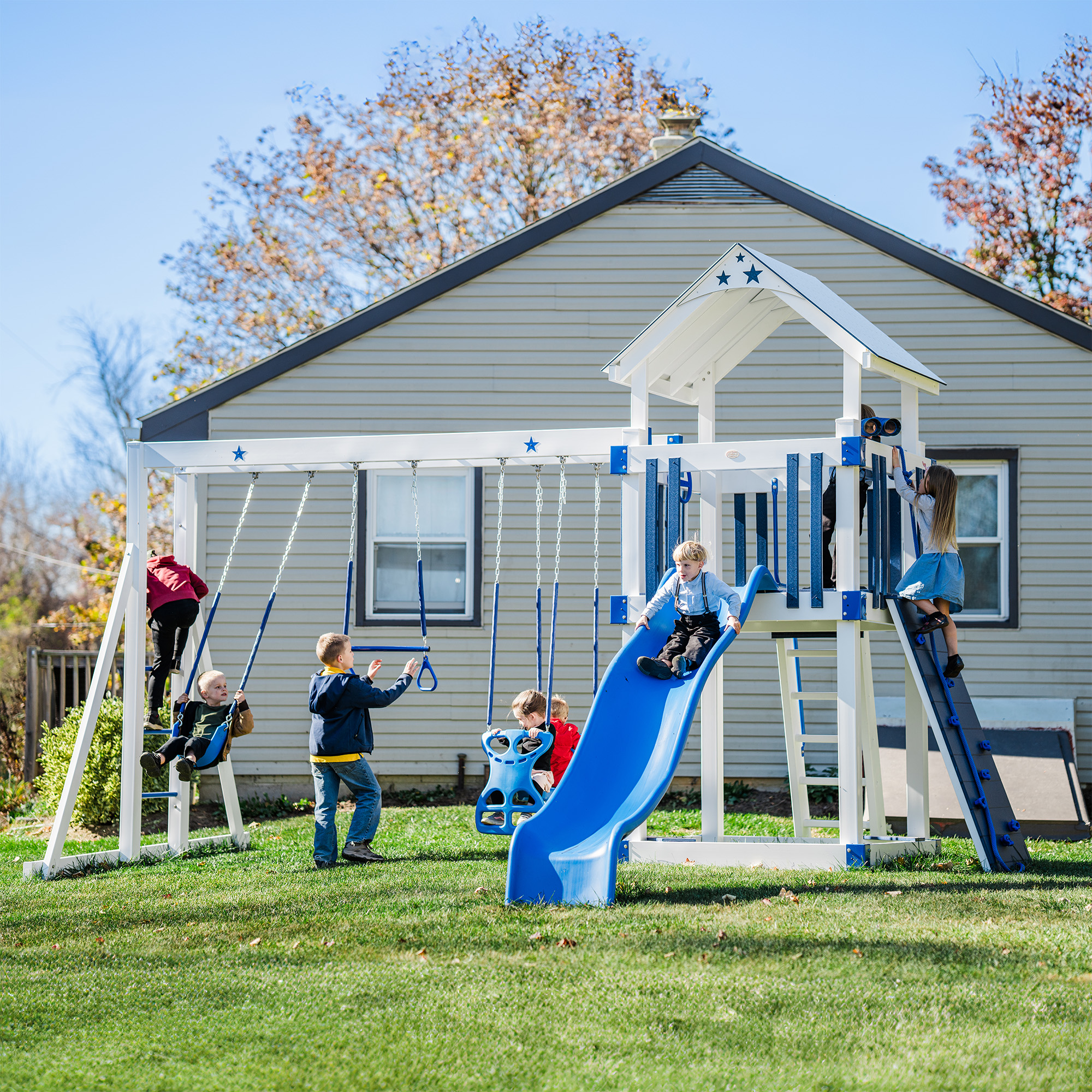 A sunny day at a playground, featuring slides, a swing set, and a small house structure in a grassy yard. Surrounded by autumn trees and a wooden fence, this idyllic scene in the heart of Toledo, OH.