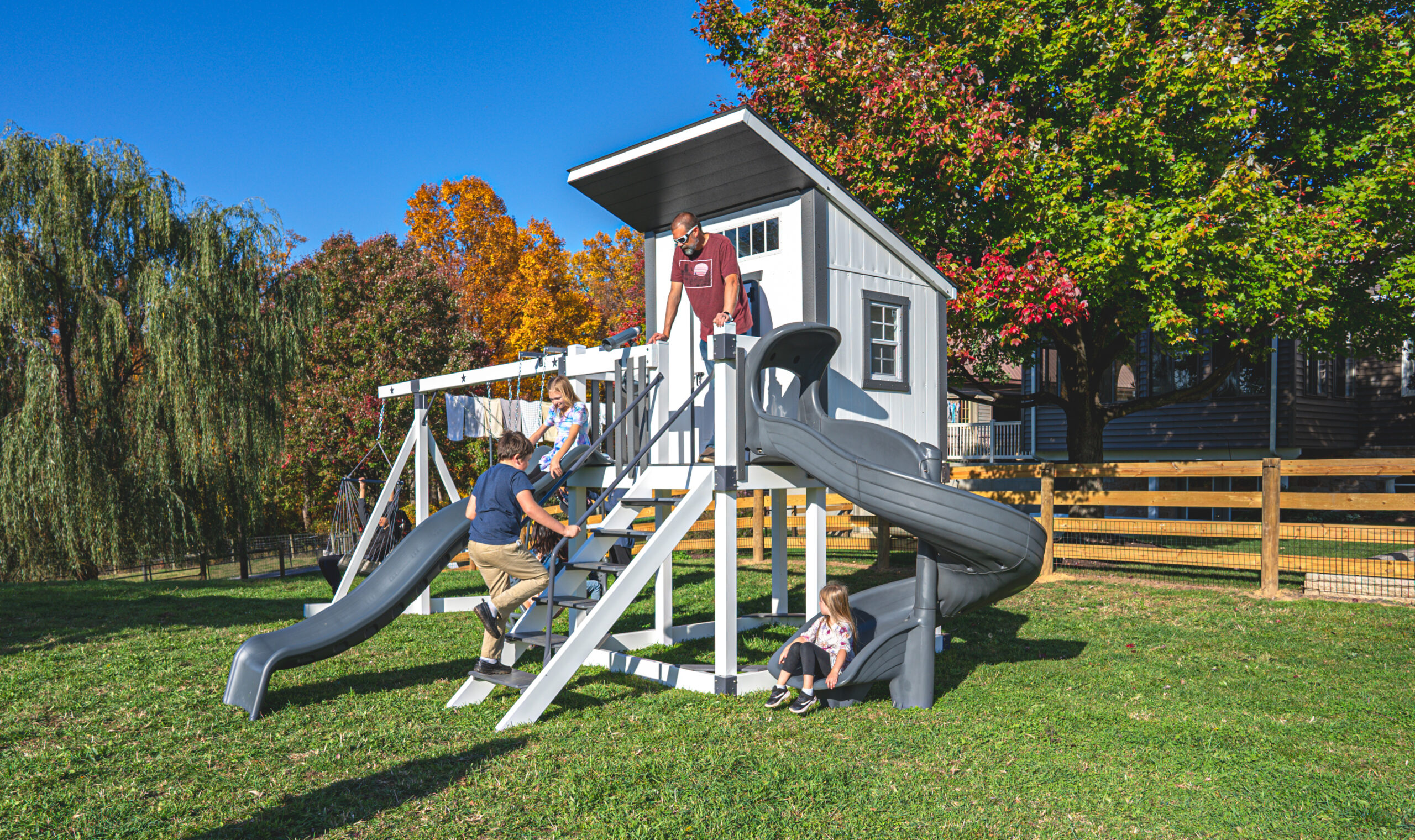 Children and an adult enjoy a sunny day at a playground, featuring slides, a swing set, and a small house structure in a grassy yard. Surrounded by autumn trees and a wooden fence, this idyllic scene in the heart of Charlotte, NC.